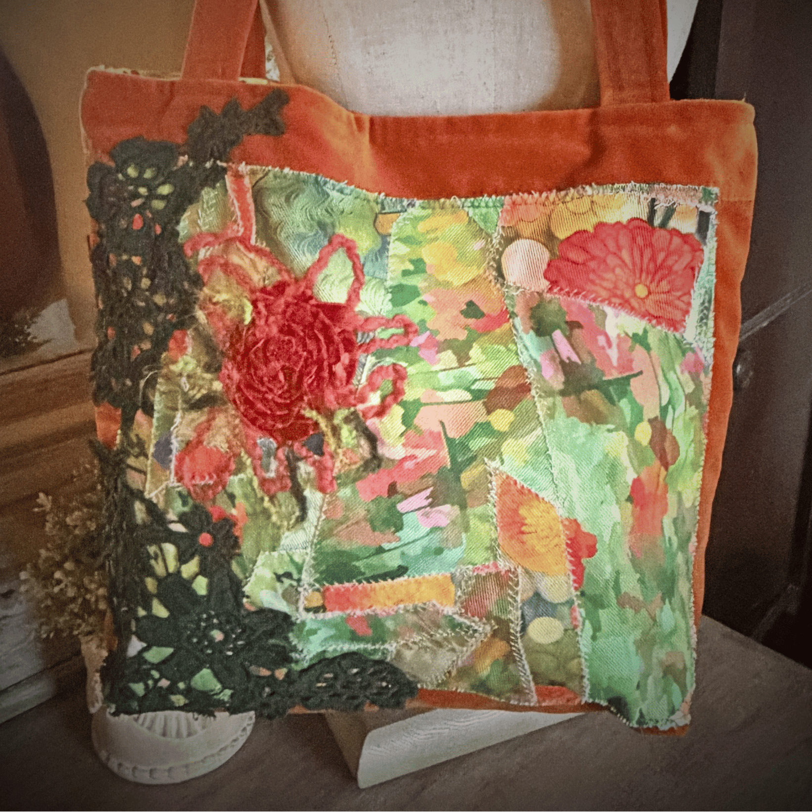 A square patchwork tote bag with orange velvet trim and handles, featuring a colorful floral and lace applique design on a green patterned fabric base, displayed on a mannequin torso.