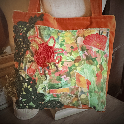 A square patchwork tote bag with orange velvet trim and handles, featuring a colorful floral and lace applique design on a green patterned fabric base, displayed on a mannequin torso.