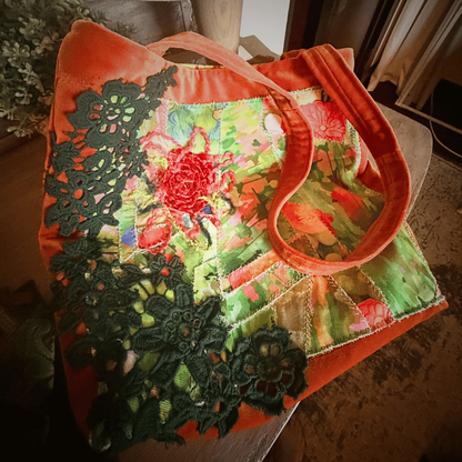 A square canvas tote bag with a vibrant floral print, featuring a bright orange border and handles, resting on a dark surface.