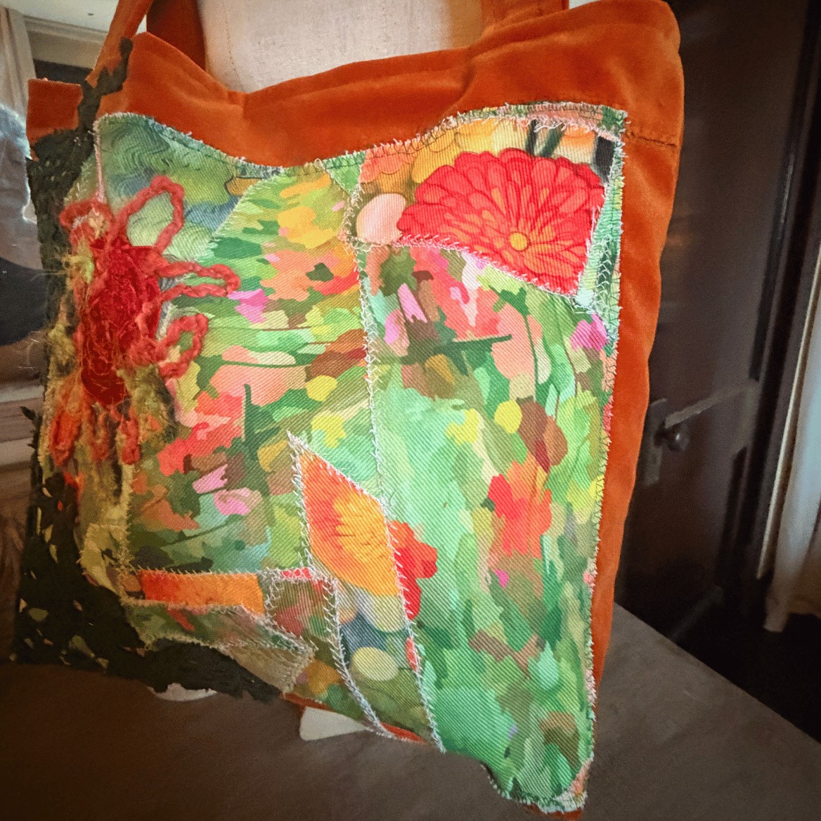 A square patchwork tote bag with a vibrant green, red, and orange floral and koi fish pattern on the front, finished with a solid orange-brown velvet trim and shoulder strap.