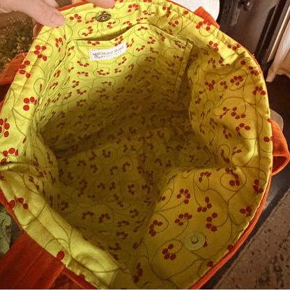 A close-up view of a quilted bag's interior lining, featuring a yellow fabric with a red cherry and green vine floral pattern, held open by a hand.